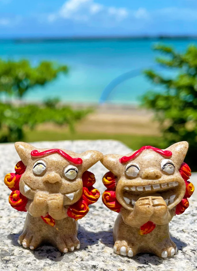 Two small beige stone shisa figurines with red manes sit on a granite ledge overlooking the ocean.