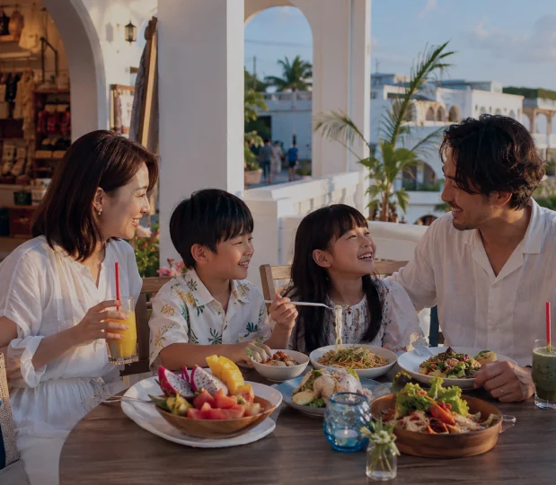A family enjoys a meal at a round wooden table on a sunlit terrace in Okinawa.