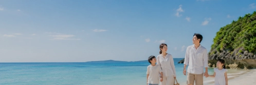 A family in light-colored clothing walks along a sandy beach beside a steep, vegetation-covered coastal cliff.