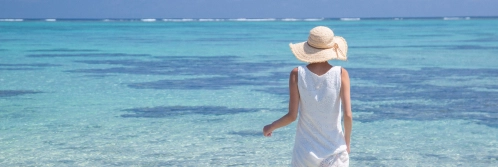 A person in a white sleeveless dress and straw hat stands before the shallow, translucent Okinawa sea.
