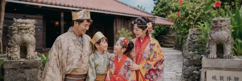 A family in vibrant patterned Ryukyu garments stands between two stone shisa statues near a wooden building.