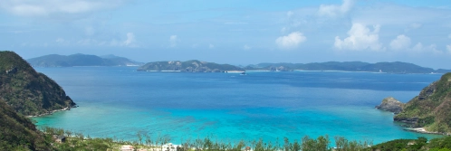 Deep blue sea transitions to shallow cyan waters between high, dark green coastal hills in Okinawa.