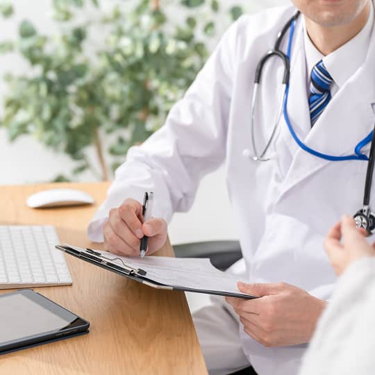 A doctor in a white coat writes on a clipboard at a light wood desk with electronics.