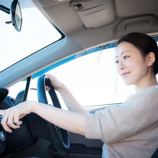 A woman in a beige knit top grips the black steering wheel inside a bright vehicle.