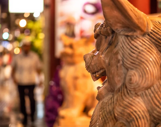 A carved wooden shisa sculpture with detailed circular mane patterns stands in profile against a blurred background.