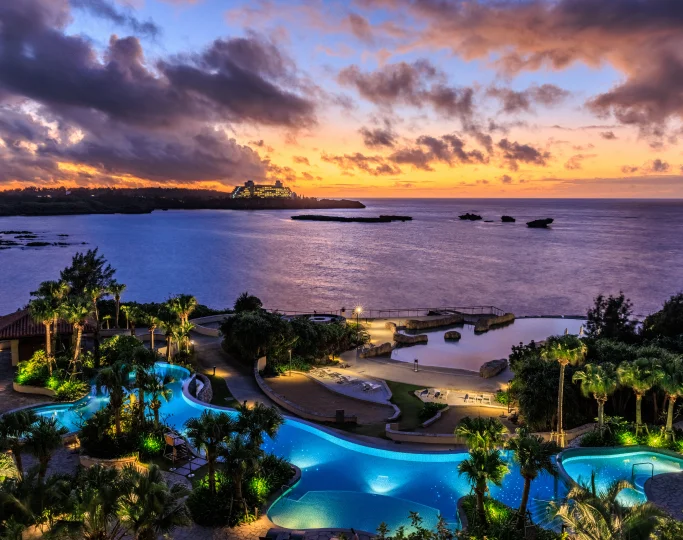 Illuminated swimming pools reflect a purple and orange sunset over the rippling sea in Okinawa.