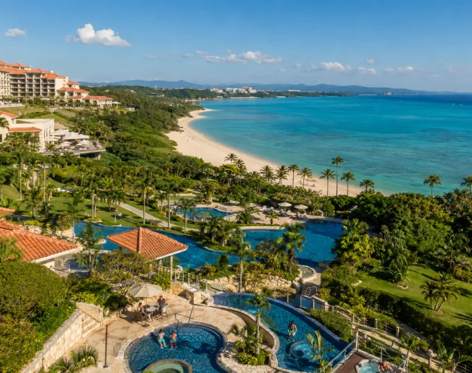 Tiered swimming pools with terracotta roofs overlook a white sand beach and coral sea in Okinawa.