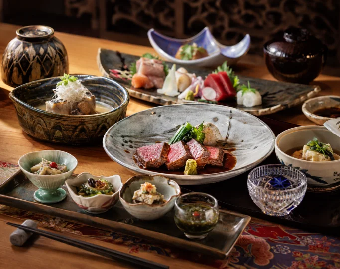 Slices of seared beef, fresh sashimi, and small side dishes are arranged on a wooden table.