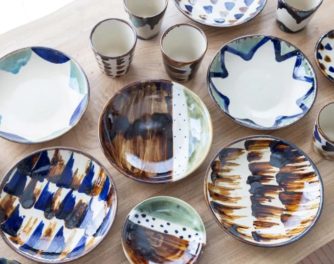 Hand-painted ceramic bowls and cups with indigo and brown brushwork sit on a light wood surface.