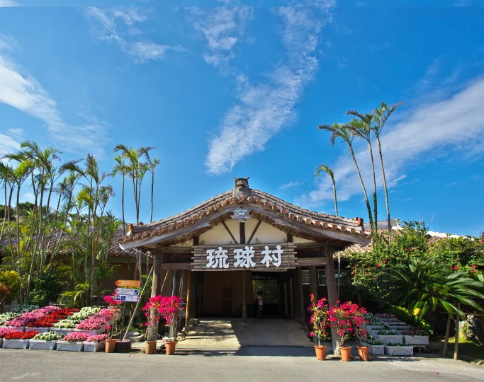 A wooden gate with traditional terracotta roof tiles stands before Ryukyu Mura amidst vibrant pink bougainvillea.